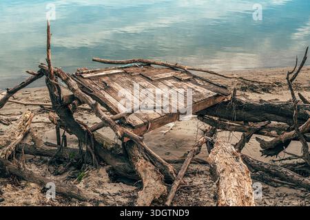 Alte verwitterte hölzerne Plattformreste kollabierten und verstrickten sich in verdrehten Treibholzzweigen auf einem Sandufer mit ruhigem Wasser im Hintergrund, se Stockfoto