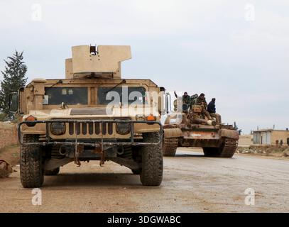 **Östliches Umland von Aleppo, Gouvernement Rakka, Syrien, Asien.** Militärfahrzeuge der syrischen Armee auf einer Landstraße, im Vordergrund gepanzertes Fahrzeug, im Hintergrund ein Panzer mit mehreren Soldaten auf dem Turm, teils mit erhobener Hand. Zivile Bebauung und Bäume am Straßenrand, bewölkter Himmel. Dokumentation militärischer Präsenz nach Gebietsgewinnen. Perspektive: Frontalansicht, leichte totale. Schlagworte: News, Politik, Sicherheit, Militär, Konflikt, Kontrolle, Symbolfoto. *** Östlicher Stadtrand von Aleppo, Gouvernement Rakka, Syrien, asiatische syrische Armee Militärfahrzeuge auf einem Can Stockfoto