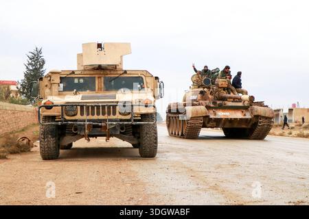 **Östliches Umland von Aleppo, Gouvernement Rakka, Syrien, Asien.** Militärfahrzeuge der syrischen Armee auf einer Landstraße, im Vordergrund gepanzertes Fahrzeug, im Hintergrund ein Panzer mit mehreren Soldaten auf dem Turm, teils mit erhobener Hand. Zivile Bebauung und Bäume am Straßenrand, bewölkter Himmel. Dokumentation militärischer Präsenz nach Gebietsgewinnen. Perspektive: Frontalansicht, leichte totale. Schlagworte: News, Politik, Sicherheit, Militär, Konflikt, Kontrolle, Symbolfoto. *** Östlicher Stadtrand von Aleppo, Gouvernement Rakka, Syrien, asiatische syrische Armee Militärfahrzeuge auf einem Can Stockfoto