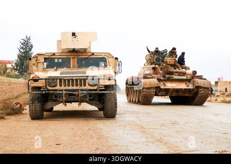 **Östliches Umland von Aleppo, Gouvernement Rakka, Syrien, Asien.** Militärfahrzeuge der syrischen Armee auf einer Landstraße, im Vordergrund gepanzertes Fahrzeug, im Hintergrund ein Panzer mit mehreren Soldaten auf dem Turm, teils mit erhobener Hand. Zivile Bebauung und Bäume am Straßenrand, bewölkter Himmel. Dokumentation militärischer Präsenz nach Gebietsgewinnen. Perspektive: Frontalansicht, leichte totale. Schlagworte: News, Politik, Sicherheit, Militär, Konflikt, Kontrolle, Symbolfoto. *** Östlicher Stadtrand von Aleppo, Gouvernement Rakka, Syrien, asiatische syrische Armee Militärfahrzeuge auf einem Can Stockfoto