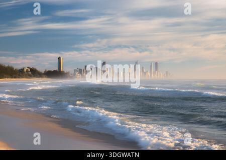 Goldenes Morgenlicht und Wellen, die am Burleigh Heads Beach, Gold Coast, Queensland mit der Skyline von Surfers Paradise ins Meer kommen. Stockfoto