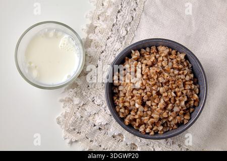 Gekochter Buchweizen in einer grauen Schüssel und ein Glas Milch auf einer Baumwolltischdecke. Stockfoto