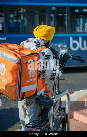 Junger asiatischer Mann mit gelbem Turban mit einer großen orangefarbenen isolierten Tasche von „JUST EAT“-Fahrern, die auf sein Handy für die nächste Bestellung schauen. Stockfoto