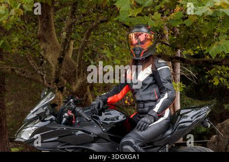Weibliche Fahrerin in Motorradanzug, Helm, Handschuhen und Stiefeln, die auf einem schwarzen Sportrad sitzt Stockfoto