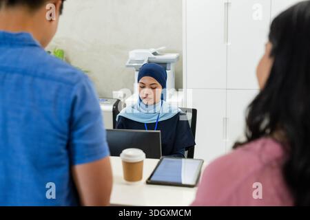Verschiedene Angestellte an der Rezeption, die blaues Kopftuch trugen und sich mit Kunden am Empfang auf einem Laptop unterhalten Stockfoto