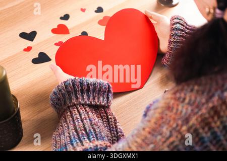Überschulteransicht einer Person in einem farbenfrohen Strickpullover, die ein großes rotes Papierherz über einem Holztisch mit Kerzen hält. Valentinstag-Konzept Stockfoto