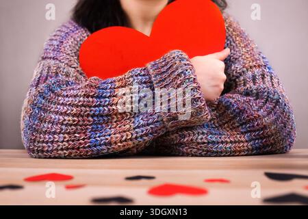 Frau in einem kuscheligen Strickpullover, der ein großes rotes Papierherz nahe der Brust umarmt. Selbstliebe, Sehnsucht oder Valentinstag-Romantik. Hohe Qualität Stockfoto
