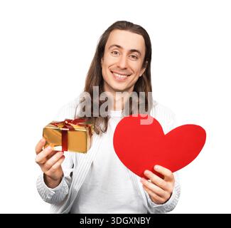 Lächelnder junger Mann mit langen Haaren, der ein rotes Papierherz und eine goldene Geschenkbox auf weißem Hintergrund hält. Romantisches Valentinstag-Konzept. Hohe qu Stockfoto