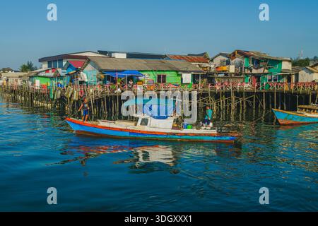 Balikpapan, Indonesien - 8. November 2025. Ein kleines, hell bemaltes traditionelles Fischerboot mit zwei Männern an Bord nähert sich einer dichten, farbenfrohen Küste Stockfoto