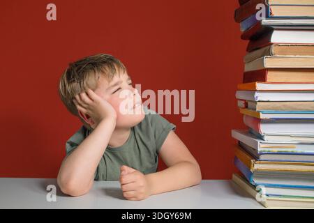 Junge und ein Stapel Bücher auf dem Tisch. Ein müder Teenager, 8-10 Jahre alt, der sich eine große Anzahl von Büchern anschaut Stockfoto