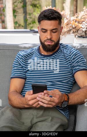 Bärtiger Mann, der auf einem grauen Sofa auf der überdachten Terrasse sitzt und ein schwarzes Smartphone hält, mit gestreiftem Hemd Stockfoto