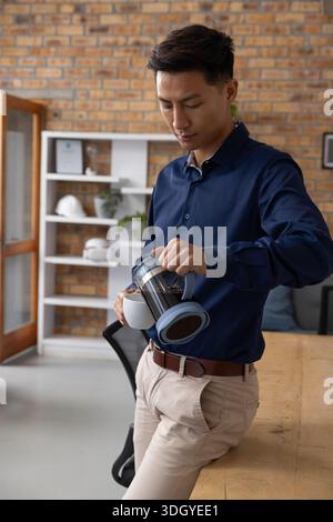 Asiatischer Mann, der Kaffee aus der französischen Presse am Bürotisch mit Helmen auf dem Regal gießt Stockfoto