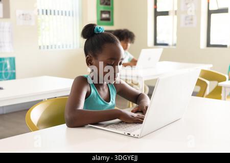 Klassenkameraden im Schulalter, die im Klassenzimmer mit weißem Laptop und gelben Stühlen auf Laptops schreiben Stockfoto