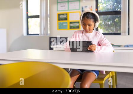 Ein junges afroamerikanisches Mädchen sitzt am Schreibtisch im Klassenzimmer und trägt Kopfhörer mit schwarzem Tablet Stockfoto