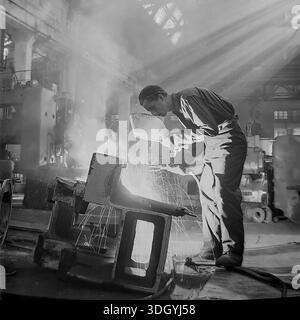 Slawjansk, Ukrainische SSR, Sowjetunion, 1975. Ein spektakuläres Archivfoto, das in der schweren Maschinenfabrik Slavtyazhmash aufgenommen wurde und einen Schweißer zeigt, der an einem massiven Stahlbauteil arbeitet. Der Arbeiter hält einen Schutzschild, während Funken aus dem Lichtbogen fliegen, was einen dramatischen Kontrast erzeugt. Die Szene wird von starken Sonnenstrahlen beleuchtet, die durch das Dach strömen und sich mit industriellem Dunst vermischen. Dieses künstlerische Bild fängt die Intensität der qualifizierten Arbeit und der Fertigung in der friedlichen Donbass-Region ein Stockfoto