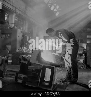 Slawjansk, Ukrainische SSR, Sowjetunion, 1975. Ein spektakuläres Archivfoto, das in der schweren Maschinenfabrik Slavtyazhmash aufgenommen wurde und einen Schweißer zeigt, der an einem massiven Stahlbauteil arbeitet. Der Arbeiter hält einen Schutzschild, während Funken aus dem Lichtbogen fliegen, was einen dramatischen Kontrast erzeugt. Die Szene wird von starken Sonnenstrahlen beleuchtet, die durch das Dach strömen und sich mit industriellem Dunst vermischen. Dieses künstlerische Bild fängt die Intensität der qualifizierten Arbeit und der Fertigung in der friedlichen Donbass-Region ein Stockfoto