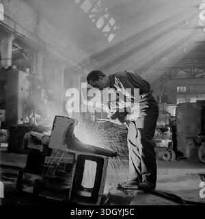 Slawjansk, Ukrainische SSR, Sowjetunion, 1975. Ein spektakuläres Archivfoto, das in der schweren Maschinenfabrik Slavtyazhmash aufgenommen wurde und einen Schweißer zeigt, der an einem massiven Stahlbauteil arbeitet. Der Arbeiter hält einen Schutzschild, während Funken aus dem Lichtbogen fliegen, was einen dramatischen Kontrast erzeugt. Die Szene wird von starken Sonnenstrahlen beleuchtet, die durch das Dach strömen und sich mit industriellem Dunst vermischen. Dieses künstlerische Bild fängt die Intensität der qualifizierten Arbeit und der Fertigung in der friedlichen Donbass-Region ein Stockfoto