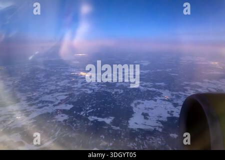 Luftaufnahme aus dem Flugzeugfenster während der Winterdämmerung. Schneebedeckte Landschaft, Stadtlichter und Motor sichtbar Stockfoto