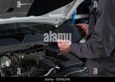 Anonymer Mann, der ein Diagnosetablett verwendet, während er den Motorraum des Autos überprüft, das elektronische Gerät mit Diagrammen und Daten hält und sich auf die Wartungsaufgabe im Fahrzeug konzentriert Stockfoto