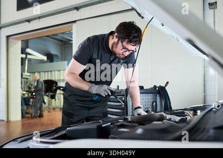 Kaukasischer junger Erwachsener, der eine Brille trägt, arbeitet an einem Automotor in einer Autowerkstatt, verwendet Werkzeug bei der Inspektion unter der Haube, konzentriert sich auf Wartungsaufgaben in der professionellen Werkstatt Stockfoto
