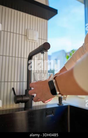 Handfüllendes Glas mit Trinkwasser aus Küchenarmatur im modernen Wohnraum. Frau gießt klares Wasser aus dem Leitungsfilter. Trinkwasserkonzept, tägliche Hydratation und Durstlöschung Stockfoto