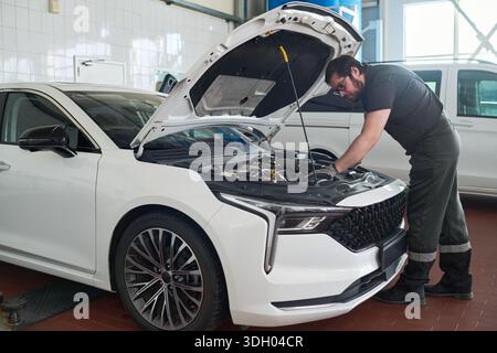 Kaukasischer Mann mittleren Alters repariert Motor unter offener Motorhaube eines modernen Autos in einer Autowerkstatt, stehend und konzentriert sich auf mechanische Arbeiten, sichtbare Werkzeuge und Fahrzeuginnenraum Stockfoto