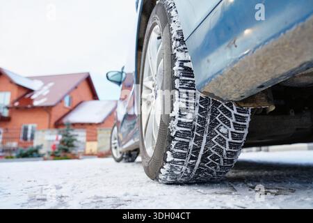 Flacher Blick auf das Autorad mit Winterreifen auf verschneite Straße in der Nähe eines Wohnhauses. Konzept für Winterfahren, Straßensicherheit, kalte Witterungsbedingungen und saisonale Fahrzeugvorbereitung Stockfoto