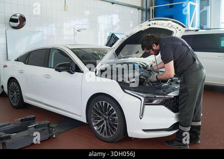 Kaukasischer junger Erwachsener, der den Motor unter der geöffneten Motorhaube einer modernen Limousine in einer Autowerkstatt repariert, neben dem Fahrzeug steht und Werkzeuge verwendet, während er sich auf mechanische Arbeiten konzentriert Stockfoto