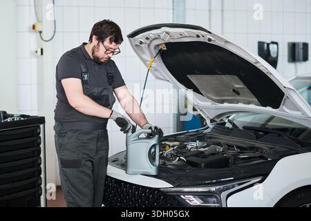 Kaukasischer junger Erwachsener, der eine Brille trägt und sich darauf vorbereitet, Motoröl in den Motor zu gießen, während er mit geöffneter Motorhaube in der Autowerkstatt steht und sich auf Wartungsaufgaben konzentriert Stockfoto