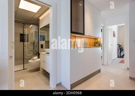 Heller Flur mit beleuchteten Regalen, eleganten Schränken und einem modernen Bad mit Glasdusche Stockfoto