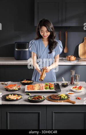 Eine junge Frau würzt gegrilltes Essen mit einer Pfeffermühle aus Holz in einer luxuriösen dunklen Küche Stockfoto