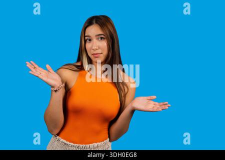 Junge Frau, die zuckte, keine Ahnung hat, sich unsicher fühlt, vor blauem Hintergrund steht Stockfoto
