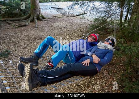 Zwei Seniorinnen entspannen in einer Hängematte in einem ruhigen Winterwald und genießen die Stille, während warme Sonnenstrahlen durch die Bäume filtern und ein friedliches A schaffen Stockfoto