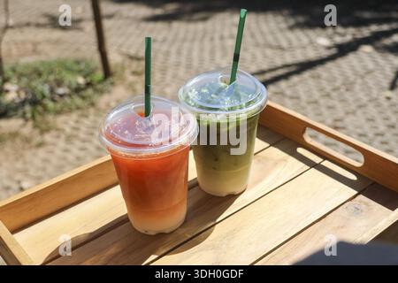Erfrischender Eistee und kalter Matcha-Latte serviert in klaren Plastikbechern mit Strohhalmen auf Holztablett für kühlen und entspannenden Sommer-Vibe im Freien Stockfoto