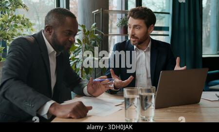 Positives Business-Team zwei multirassische Männer sprechen sorglos im Büro aussehende Laptop Geschäftsleute Firmenpräsentation Brainstorming lächelnder Kaukasier Stockfoto