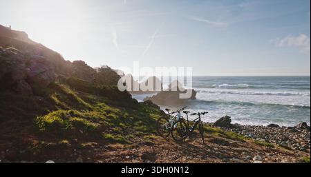 Portugal, Lissabon: Zwei Mountainbikes parken auf dem Küstenweg über felsigen Klippen, blaue Wellen des Atlantiks, sonnendurchflutetes grünes Gras und Linsenflecken wecken Sommerradfreiheit und Abenteuer Stockfoto