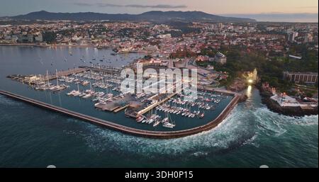 Portugal, Lissabon: Die Küstenstadt Cascais mit einer lebhaften Marina-Bucht mit Bootsbooten, historischen Gebäuden und einer weiten Stadtlandschaft, die sich in der Abenddämmerung in die Berge ausdehnt. Panoramablick aus der Vogelperspektive Stockfoto