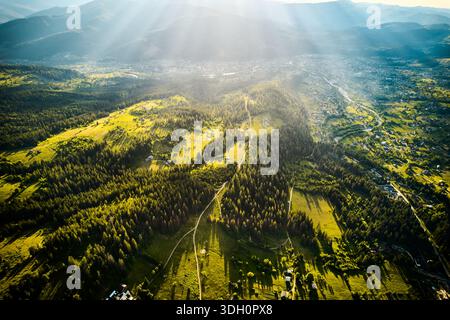 Atemberaubender Blick aus der Luft auf das Tal, im Sonnenlicht getaucht, mit Waldstücken und offenen Feldern. Verwinkelte Straßen und verstreute Häuser, umgeben von sanften Hügeln und fernen Bergen. Stockfoto