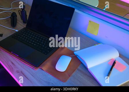 Tischeinrichtung mit Laptop, Maus, Notebook und Stift, beleuchtet durch bunte Neonlichter Stockfoto