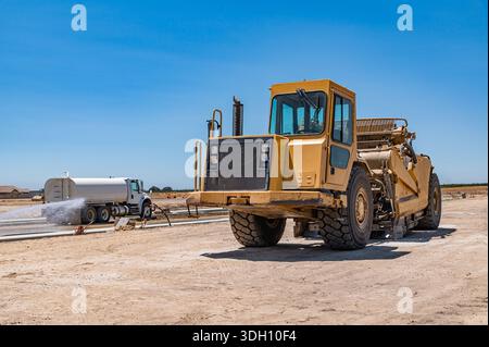 Gelber Traktor, Straßenreparatur. Seitenansicht. Bewässerungsmaschine im Hintergrund. Stockfoto