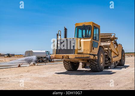 Gelber Traktor, Straßenreparatur. Seitenansicht. Bewässerungsmaschine im Hintergrund. Stockfoto