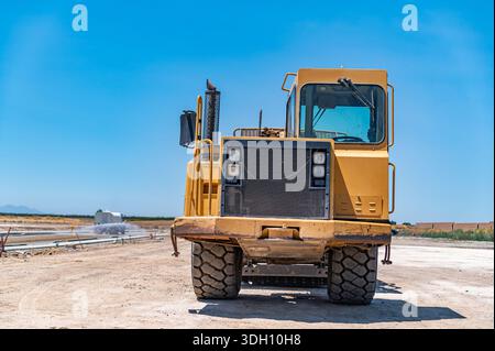 Gelber Traktor, Straßenreparatur. Nahaufnahme. Bewässerungsmaschine im Hintergrund. Stockfoto