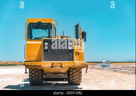 Gelber Traktor, Straßenreparatur. Nahaufnahme. Bewässerungsmaschine im Hintergrund. Stockfoto