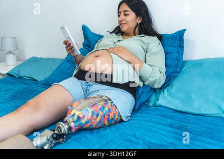 Schwangere Frau mit einer Beinprothese benutzt ihr Smartphone, während sie sich im Bett entspannt Stockfoto