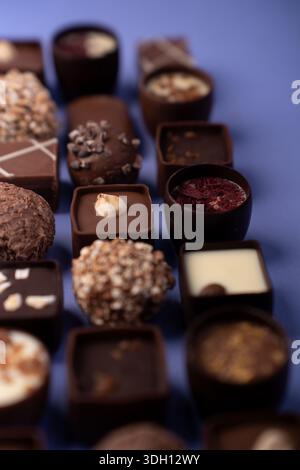 Makroaufnahme von verschiedenen Gourmet-Pralinen und Trüffeln, die in ordentlichen Reihen auf einer lila Oberfläche angeordnet sind und kräftige Brauntöne, vielfältige Texturen, Stockfoto