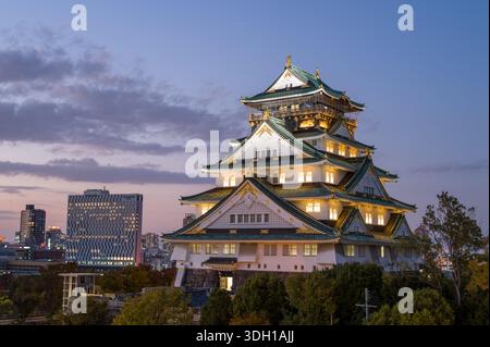 Osaka, Japan - 7. November 2023: Der Hauptturm der Burg Osaka wird in der Abenddämmerung beleuchtet, mit modernen Stadtgebäuden und Bäumen im Hintergrund unter teilweise bewölktem Himmel. Stockfoto