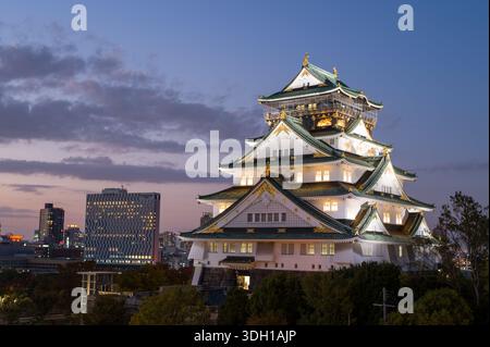 Osaka, Japan - 7. November 2023: Der Hauptturm der Burg Osaka wird in der Abenddämmerung beleuchtet, mit modernen Stadtgebäuden und Bäumen im Hintergrund unter teilweise bewölktem Himmel. Stockfoto