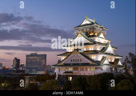 Osaka, Japan - 7. November 2023: Der Hauptturm der Burg Osaka wird bei Nacht beleuchtet, mit modernen Stadtgebäuden und Bäumen im Hintergrund unter dem Dämmerungshimmel. Stockfoto