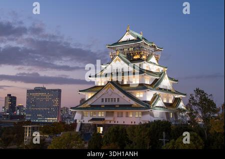 Osaka, Japan - 7. November 2023: Der Hauptturm der Burg Osaka wird in der Abenddämmerung beleuchtet, mit modernen Stadtgebäuden und Bäumen im Hintergrund unter teilweise bewölktem Himmel. Stockfoto