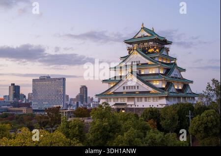 Osaka, Japan - 7. November 2023: Der Hauptturm der Burg Osaka wird in der Abenddämmerung beleuchtet, mit modernen Stadtgebäuden und Bäumen im Hintergrund unter teilweise bewölktem Himmel. Stockfoto